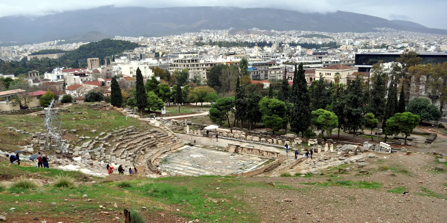 A wide view of the Theatre of Dionysus below the Acropolis wall, with the city of Athens behind it.