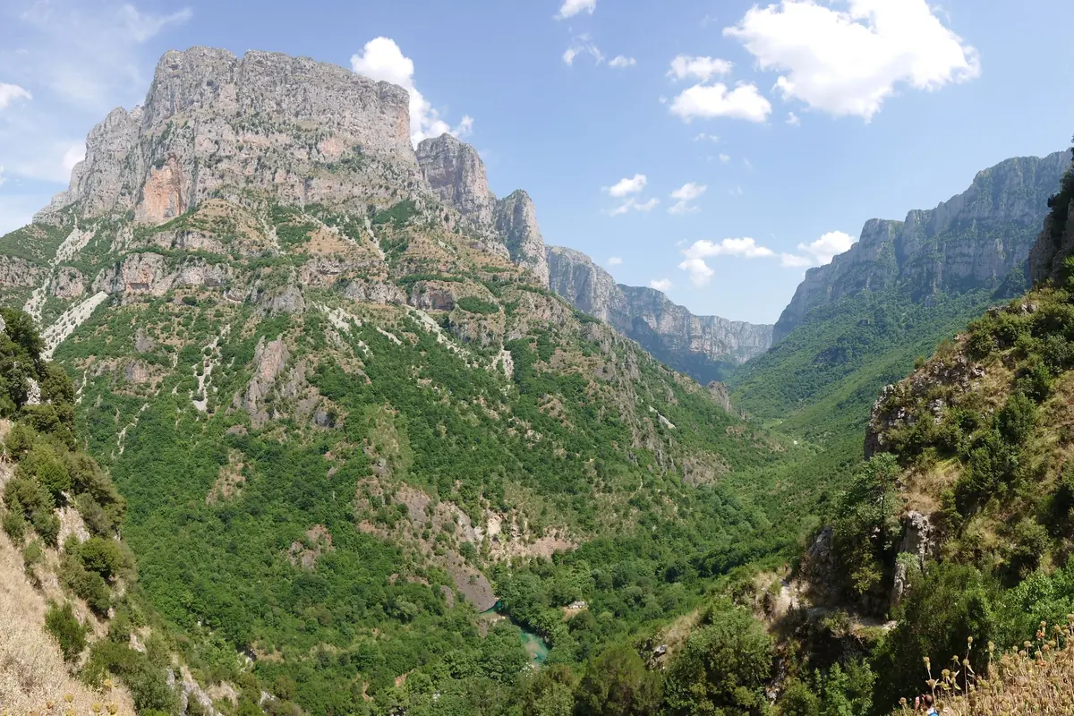 A deep green gorge below tall limestone cliffs in Vikos Gorge, with a blue sky above.