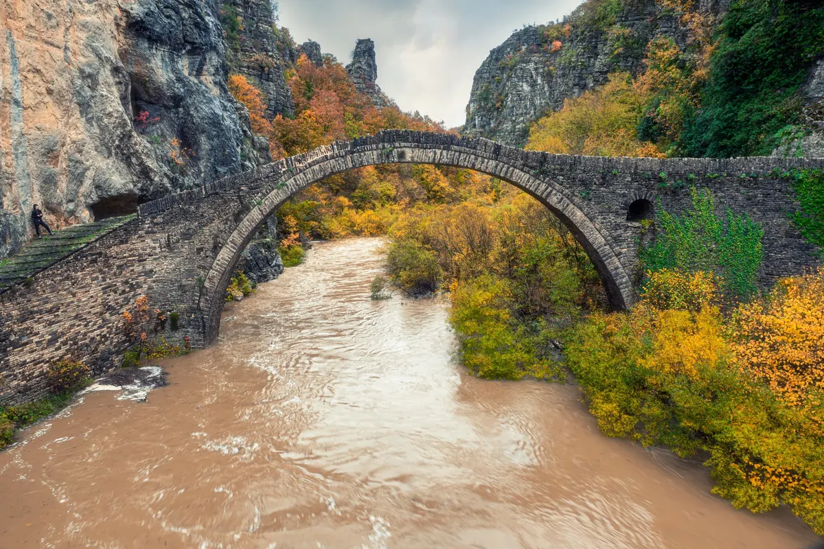 An old stone arch bridge crossing a brown river in autumn colours, between rocky slopes in the Pindus mountains of northern Greece.