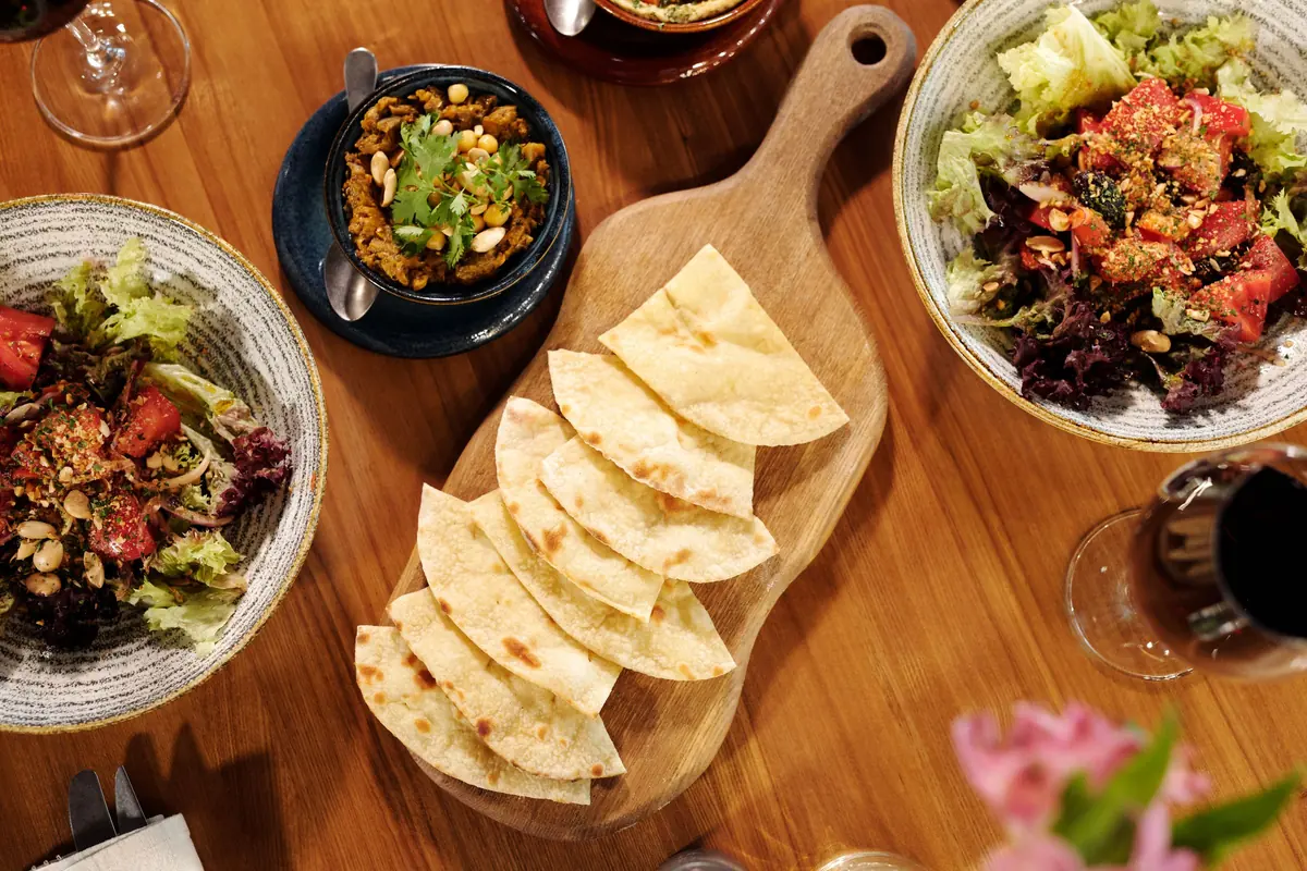 A wooden table from above, with salads, flatbread slices on a board, a small bowl of food and glasses of wine.
